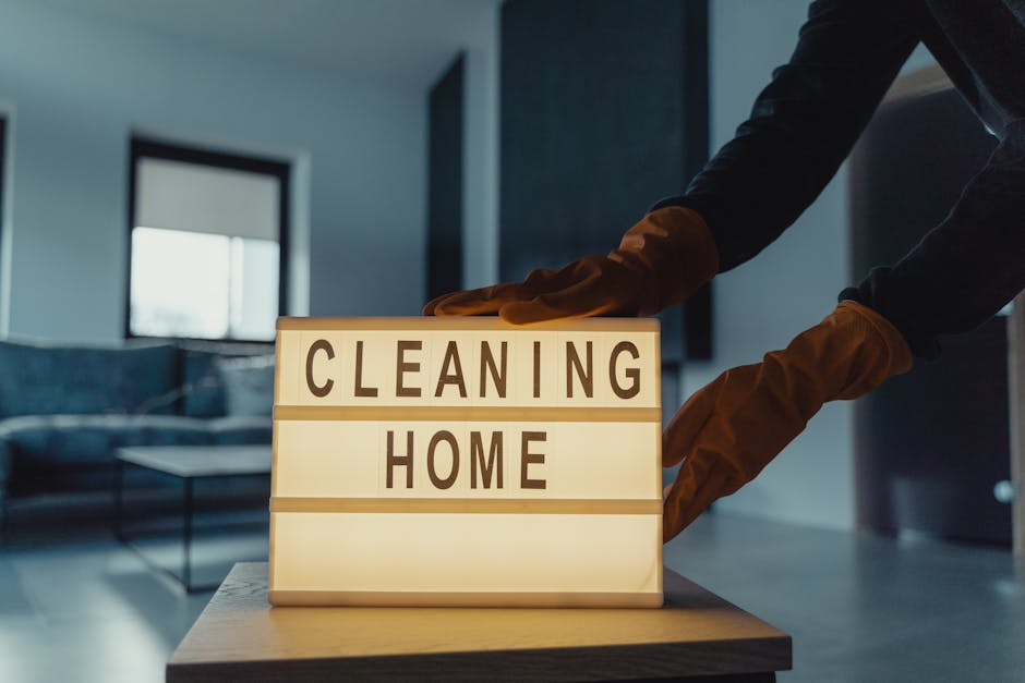 In a modern living room with a dark-colored sofa, a person wearing black sleeves and brown gloves is placing a lit sign on a wooden table. The sign displays the words 'CLEANING HOME' in bold capital l